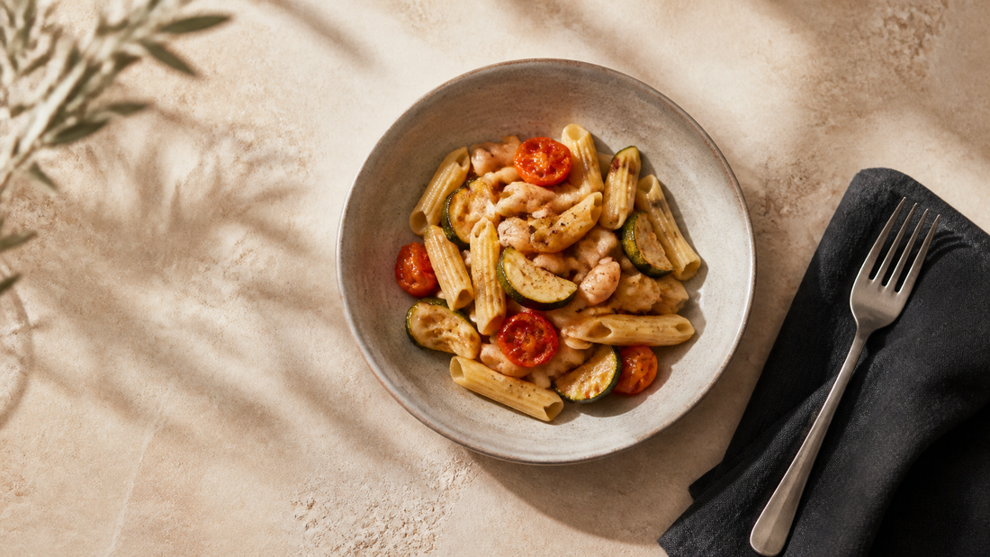 Cuenco de pasta corta con calabacín, tomate y alubias junto a un tenedor sobre una mesa clara.